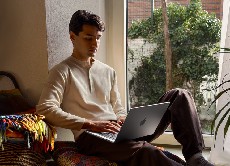 Une personne assise par terre à côté d’une fenêtre utilisant un MacBook.