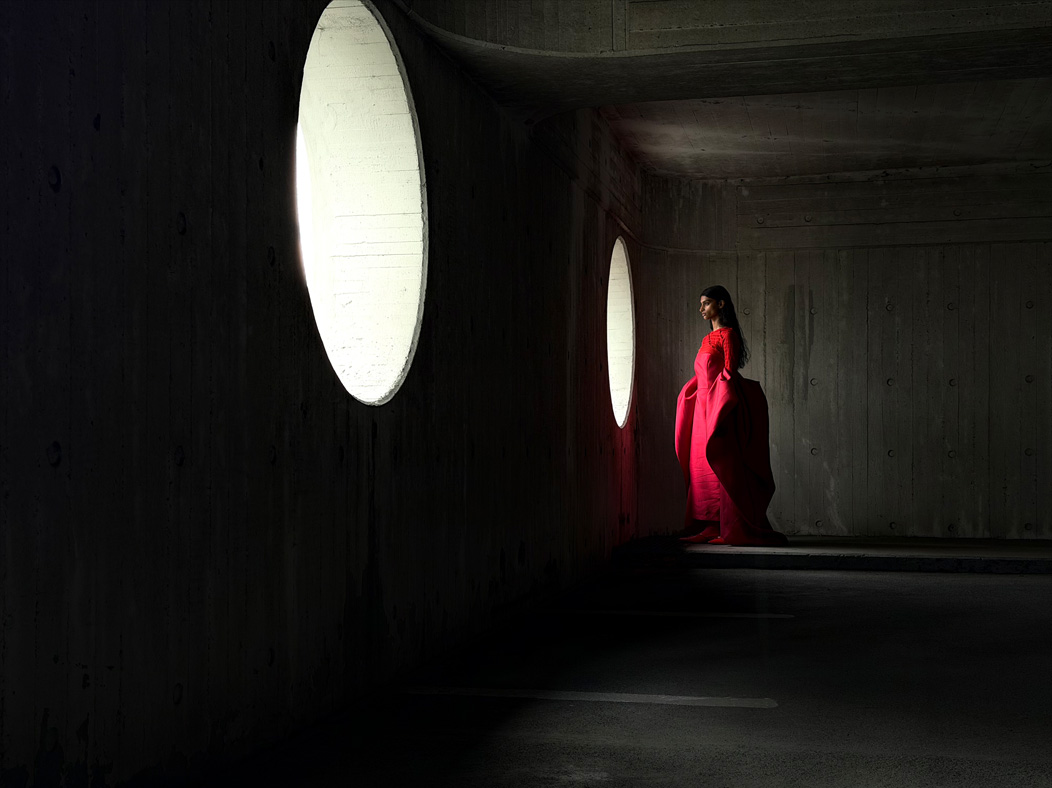 Una mujer con un largo vestido rojo de pie en medio de una habitación vacía mientras la fotografían. La distancia focal de la imagen se va alternando en la app Fotos para mostrar las capacidades del sistema de cámaras Pro: 48 mm